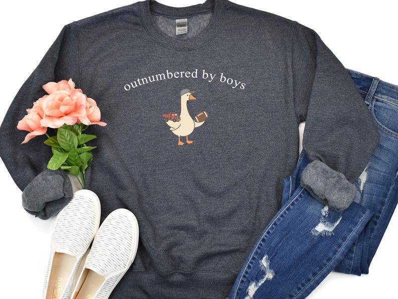 May include: Dark gray sweatshirt with the text "outnumbered by boys" above a cartoon goose wearing a hat and holding a football. The sweatshirt is paired with blue jeans and white shoes.