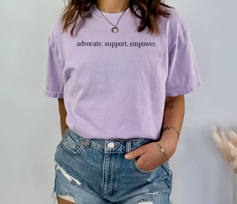 Op de afbeelding: Een lichtpaars t-shirt met de woorden "advocate. support. empower." in zwarte letters. Het shirt wordt gecombineerd met een spijkerbroek. De stijl is casual en comfortabel.