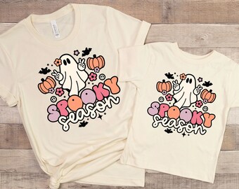 May include: Two cream colored t-shirts with a ghost, pumpkins, and flowers graphic. The text "Spooky Season" is in pink, orange, and purple letters.