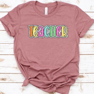 Puede incluir: Una camiseta color malva con la palabra "teacher" impresa en la parte delantera en letras de colores con un patr&oacute;n de lunares blancos y negros.