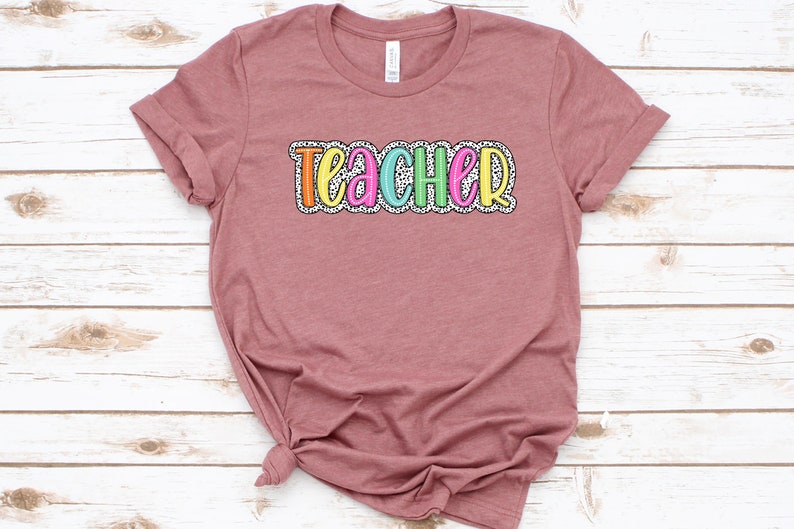 Puede incluir: Una camiseta color malva con la palabra "teacher" impresa en la parte delantera en letras de colores con un patr&oacute;n de lunares blancos y negros.