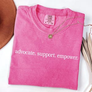 Op de afbeelding: Een felroze t-shirt met de witte tekst "advocate. support. empower.". Een gouden ketting met een hanger ligt op het shirt. Het shirt is opgevouwen en heeft een Comfort Colors label.