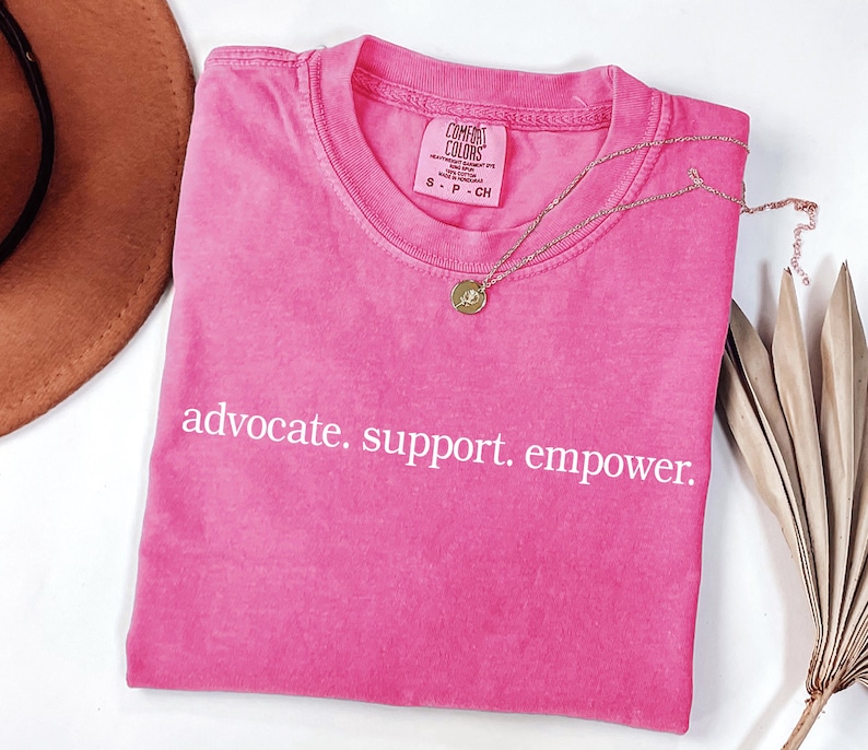 Op de afbeelding: Een felroze t-shirt met de witte tekst "advocate. support. empower.". Een gouden ketting met een hanger ligt op het shirt. Het shirt is opgevouwen en heeft een Comfort Colors label.