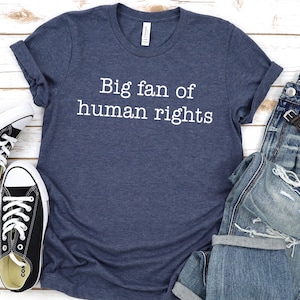 May include: A navy blue t-shirt with the text "Big fan of human rights" in white, paired with distressed blue jeans, a wristwatch, and black and white sneakers. The items are arranged on a light wood-grain surface.