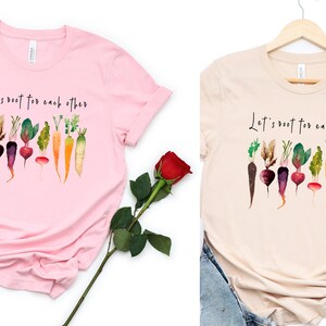 May include: Two t-shirts with a watercolor illustration of vegetables and the text "Let's root for each other". The shirts are pink and beige.