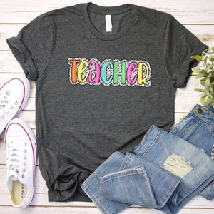 Puede incluir: Una camiseta gris oscuro con la palabra "TEACHER" en letras de colores con lunares blancos. Las letras est&aacute;n delineadas en negro.