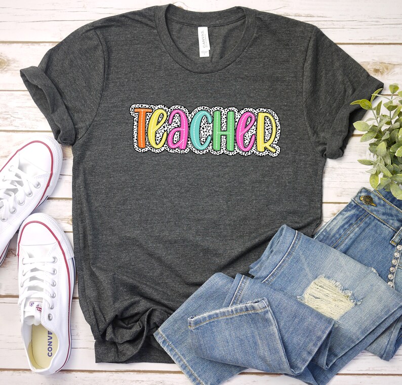 Puede incluir: Una camiseta gris oscuro con la palabra "TEACHER" en letras de colores con lunares blancos. Las letras est&aacute;n delineadas en negro.