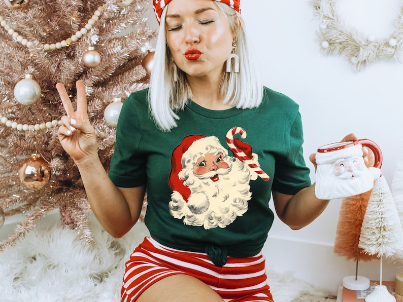 May include: A green t-shirt featuring a vintage illustration of Santa Claus holding a candy cane. The shirt is being worn by a person with blonde hair and red lipstick. The person is holding a white mug with a Santa Claus design.