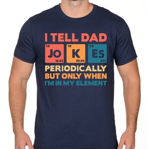 Puede incluir: Camiseta azul marino con el texto "I TELL DAD JOKES PERIODICALLY BUT ONLY WHEN I'M IN MY ELEMENT" en un dise&ntilde;o colorido de tabla peri&oacute;dica. La camiseta presenta los elementos Jo, K y Es. Una camiseta divertida con tem&aacute;tica cient&iacute;fica.