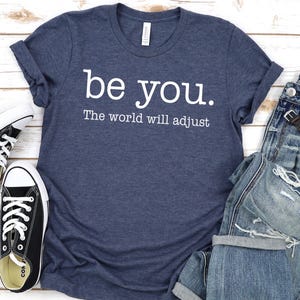 Puede incluir: Una camiseta azul marino con el texto "be you. The world will adjust" en blanco, combinada con vaqueros azules desgastados, zapatillas negras y un reloj de pulsera. El atuendo informal est&aacute; dispuesto sobre una superficie de madera clara.