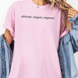 Op de afbeelding: Een lichtroze t-shirt met de zwarte tekst "advocate. support. empower.". Het t-shirt wordt gedragen met een spijkerjasje.