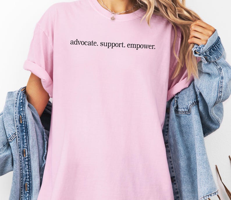 Op de afbeelding: Een lichtroze t-shirt met de zwarte tekst "advocate. support. empower.". Het t-shirt wordt gedragen met een spijkerjasje.
