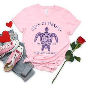Puede incluir: Una camiseta rosa claro con un dise&ntilde;o de tortuga marina azul marino y el texto "GULF OF MEXICO EST. 1550 STAY SALTY AND RESIST". La camiseta se combina con zapatillas rosas, vaqueros y una rosa roja.