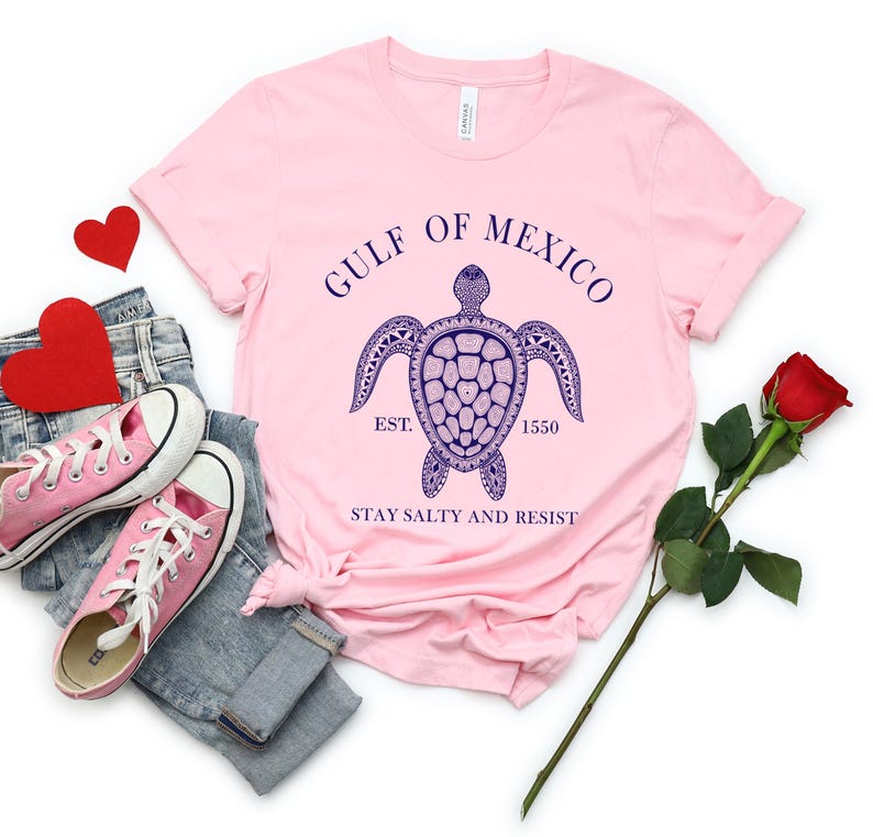 Puede incluir: Una camiseta rosa claro con un dise&ntilde;o de tortuga marina azul marino y el texto "GULF OF MEXICO EST. 1550 STAY SALTY AND RESIST". La camiseta se combina con zapatillas rosas, vaqueros y una rosa roja.