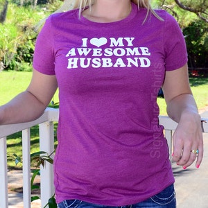 May include: A purple t-shirt with the text "I ❤️ MY AWESOME HUSBAND" in white. The shirt has short sleeves and a crew neck. The image is taken outdoors.