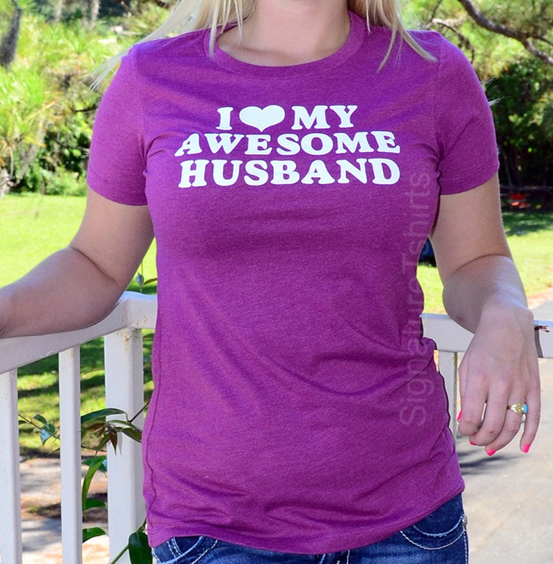 May include: A purple t-shirt with the text "I ❤️ MY AWESOME HUSBAND" in white. The shirt has short sleeves and a crew neck. The image is taken outdoors.