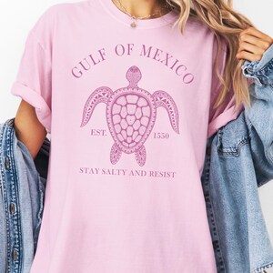 Op de afbeelding: Roze T-shirt met een afbeelding van een zeeschildpad en de tekst "Gulf of Mexico Est. 1550 Stay Salty and Resist".
