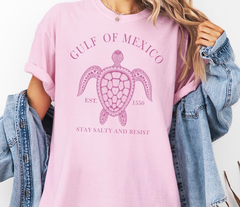 Op de afbeelding: Roze T-shirt met een afbeelding van een zeeschildpad en de tekst "Gulf of Mexico Est. 1550 Stay Salty and Resist".