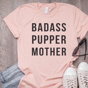 May include: A pink t-shirt with the text "BADASS PUPPER MOTHER" printed in black.