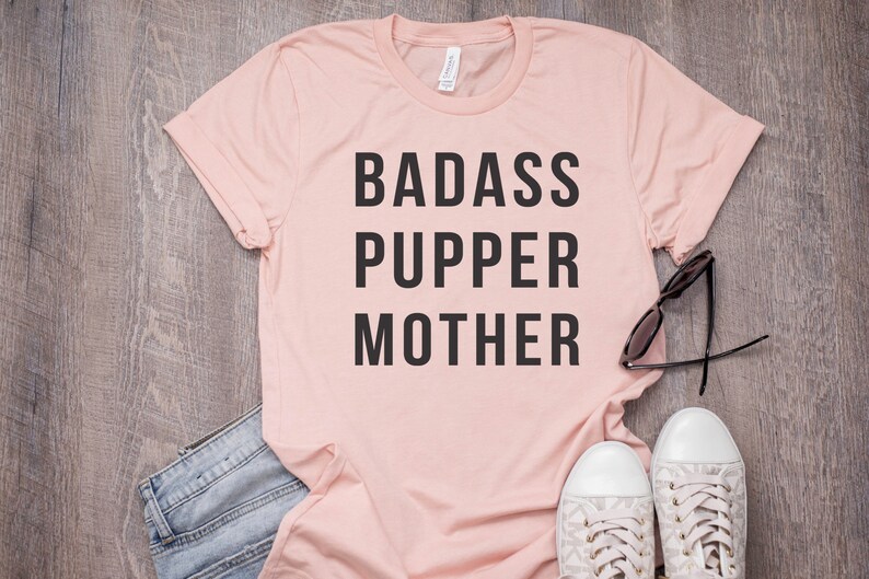 May include: A pink t-shirt with the text "BADASS PUPPER MOTHER" printed in black.
