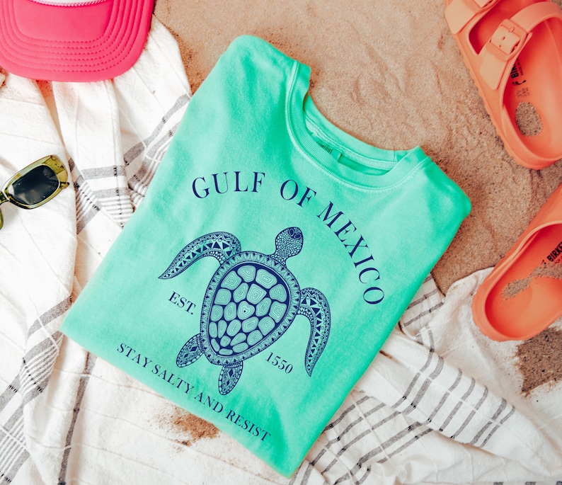 Op de afbeelding: Een turquoise t-shirt met een afbeelding van een zeeschildpad en de tekst "Gulf of Mexico Est. 1550 Stay Salty and Resist".