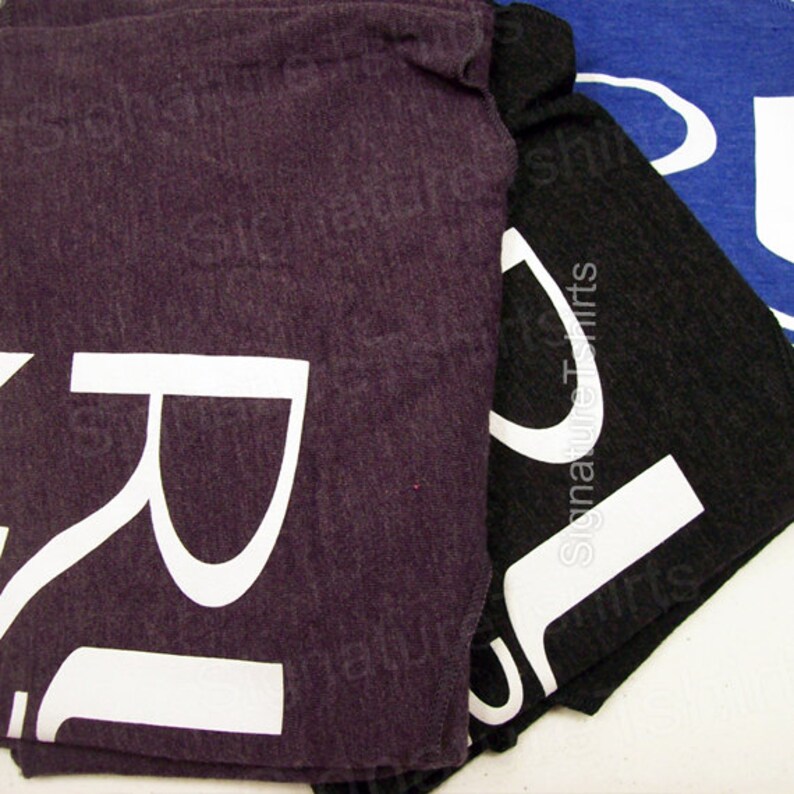May include: Three folded t-shirts with the letter "R" printed in white on each. The shirts are in different colors: purple, black, and blue.