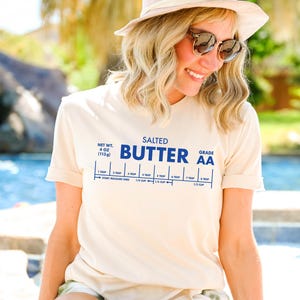May include: A white t-shirt with a blue graphic of a stick of butter with the text "SALTED BUTTER GRADE AA" and a measurement guide for tablespoons and cups. The t-shirt is being worn by a person.