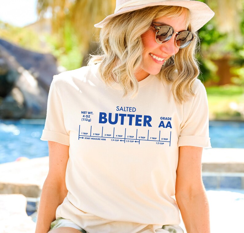 May include: A white t-shirt with a blue graphic of a stick of butter with the text "SALTED BUTTER GRADE AA" and a measurement guide for tablespoons and cups. The t-shirt is being worn by a person.