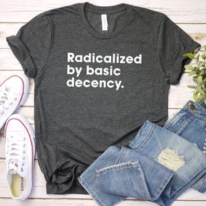 May include: A dark gray t-shirt with the text "Radicalized by basic decency." printed in white on the front. The shirt is laying on a wooden surface with a pair of white sneakers and blue jeans.