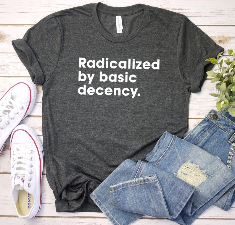 May include: A dark gray t-shirt with the text "Radicalized by basic decency" in white. The shirt is paired with folded blue jeans and white canvas sneakers with red trim.