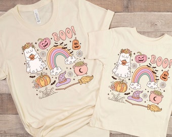 May include: Two cream-colored t-shirts with a Halloween-themed graphic. The design includes a ghost, pumpkins, a rainbow, a witch's hat, and the word "BOO!". The shirts are short-sleeved and made of a soft material.
