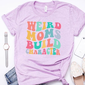 Pode incluir: Uma camiseta roxa com o texto "Weird Moms Build Character" em cores do arco-&iacute;ris. O texto est&aacute; em um estilo de fonte retr&ocirc;.