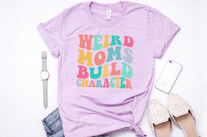 Pode incluir: Uma camiseta roxa com o texto "Weird Moms Build Character" em cores do arco-&iacute;ris. O texto est&aacute; em um estilo de fonte retr&ocirc;.