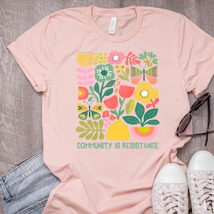 Op de afbeelding: Een roze T-shirt met een kleurrijk bloemen- en vlinderontwerp. De tekst "Community is Resistance" staat op de voorkant van het shirt.