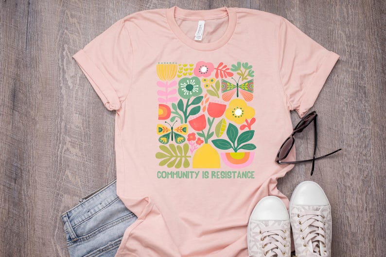 Op de afbeelding: Een roze T-shirt met een kleurrijk bloemen- en vlinderontwerp. De tekst "Community is Resistance" staat op de voorkant van het shirt.