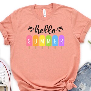 May include: A peach colored t-shirt with the text "hello summer" in black lettering. The text is surrounded by six colorful popsicle graphics.