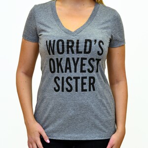 May include: A heather gray v-neck t-shirt with the text "WORLD'S OKAYEST SISTER" printed in black. The shirt is made of a soft material and has short sleeves. The text is centered on the front of the shirt.