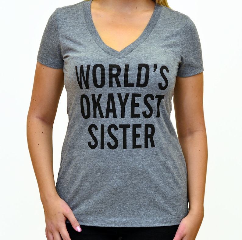 May include: A heather gray v-neck t-shirt with the text "WORLD'S OKAYEST SISTER" printed in black. The shirt is made of a soft material and has short sleeves. The text is centered on the front of the shirt.