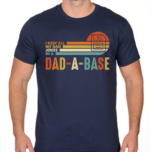 Op de afbeelding: Marineblauw T-shirt met de tekst "I KEEP ALL MY DAD JOKES IN A DAD-A-BASE" in een retro-ontwerp. De afbeelding bevat een gestileerde afbeelding van een diskette. Het T-shirt heeft een ronde hals.