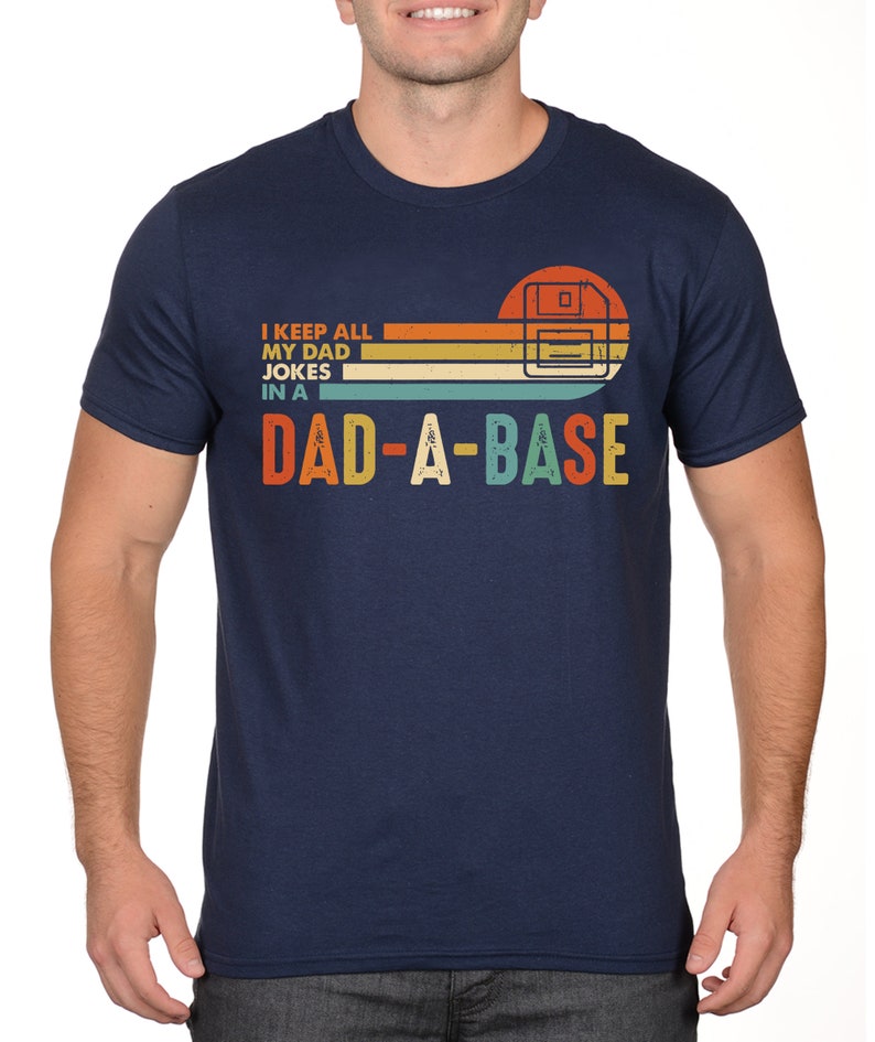 Op de afbeelding: Marineblauw T-shirt met de tekst "I KEEP ALL MY DAD JOKES IN A DAD-A-BASE" in een retro-ontwerp. De afbeelding bevat een gestileerde afbeelding van een diskette. Het T-shirt heeft een ronde hals.
