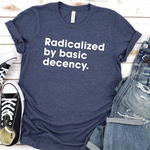 May include: A navy blue t-shirt with the text "Radicalized by basic decency" in white, paired with distressed blue jeans, black sneakers, and a wristwatch. The items are arranged on a light wood surface.