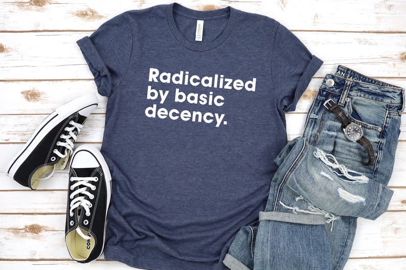 May include: A navy blue t-shirt with the text "Radicalized by basic decency" in white, paired with distressed blue jeans, black sneakers, and a wristwatch. The items are arranged on a light wood surface.