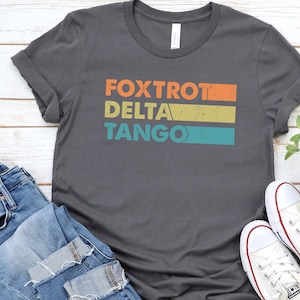 以下が含まれることがあります： オレンジ、イエロー、ブルーで「FOXTROT DELTA TANGO」と書かれたヴィンテージスタイルのグラフィックが施されたダークグレーのTシャツです。