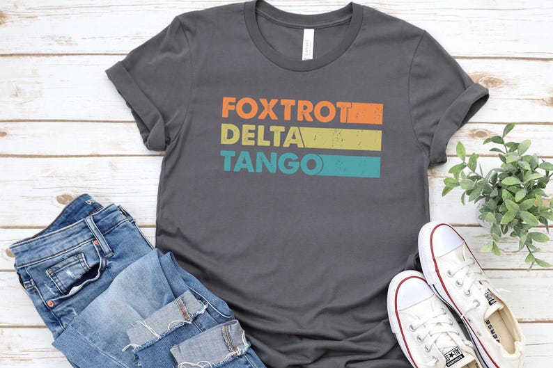 以下が含まれることがあります： オレンジ、イエロー、ブルーで「FOXTROT DELTA TANGO」と書かれたヴィンテージスタイルのグラフィックが施されたダークグレーのTシャツです。