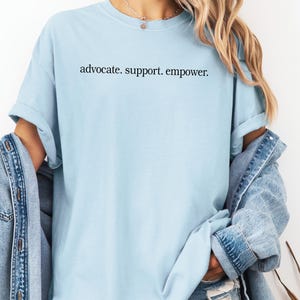 Op de afbeelding: Lichtblauw T-shirt met de zwarte tekst "advocate. support. empower.". Het shirt heeft korte mouwen en een ronde hals. Een spijkerjasje wordt over de schouders gedragen.