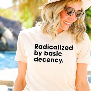 May include: A cream-colored t-shirt with the text "Radicalized by basic decency" in black. The shirt has short sleeves and is being worn outdoors. The background includes a pool and greenery.