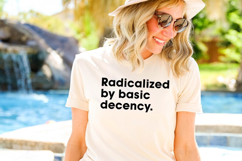 May include: A cream-colored t-shirt with the text "Radicalized by basic decency" in black. The shirt has short sleeves and is being worn outdoors. The background includes a pool and greenery.
