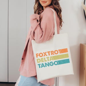以下が含まれることがあります： 「FOXTROT DELTA TANGO」とカラフルなグラフィックデザインが施された白いトートバッグです。