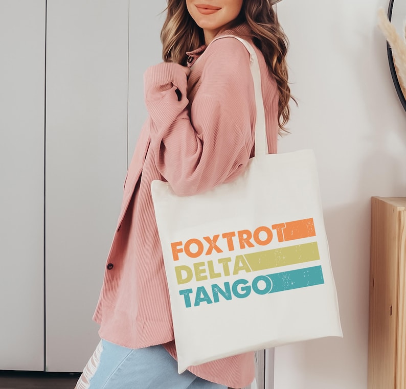 以下が含まれることがあります： 「FOXTROT DELTA TANGO」とカラフルなグラフィックデザインが施された白いトートバッグです。
