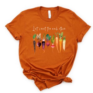 May include: Orange short-sleeved t-shirt with the text "Let's root for each other" and a watercolor illustration of various vegetables, including carrots, beets, and turnips.